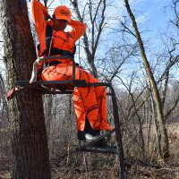 A person wearing orange sitting in a tree stand in the woods.